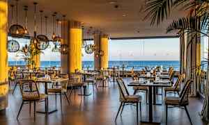 Billede af en restaurant med kurvestole, bordene er dækket med udsigt over havet gennem store glasvinduer. (AI-genereret tekst)