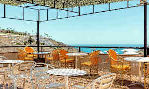 Udendørs servering med borde og stole på en terrasse med udsigt over havet og en skråning med bygninger. (AI-genereret tekst)