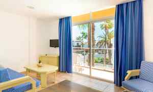 Et lyst hotelværelse med en blåstribet sofa, et bord med en flaske champagne og glas, et lille TV på en kommode og store vinduer med blå gardiner, der fører ud til en balkon med udsigt over palmer. (AI-genereret tekst)