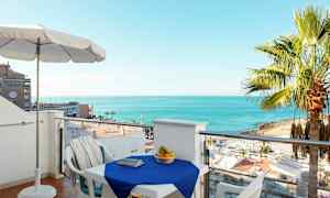 Udsigt fra en altan med et bord dækket med en blå dug, omgivet af stole. En kurv med frugt står på bordet, og i baggrunden ses stranden, palmer og havet. (AI-genereret tekst)