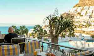 En person sidder på en terrasse med udsigt over havet, træer og bygninger, med en kop kaffe og morgenmad foran sig. (AI-genereret tekst)