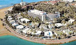 Luftfoto af en kystby med hotel, palmer og strand. (AI-genereret tekst)