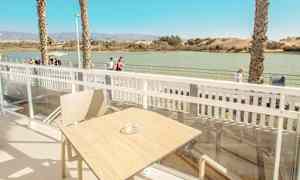 Udsigt fra en terrasse med bord og stol, med vand og gangsti i baggrunden. (AI-genereret tekst)