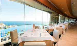 En restaurant med dækkede borde og udsigt over havet gennem store glasvinduer. (AI-genereret tekst)