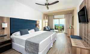 Et hotelværelse med to senge, et siddeområde og en balkon med udsigt over havet. (AI-genereret tekst)