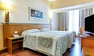 Et hotelværelse med en dobbeltseng med mønstrede sengetæpper, lamper på væggen og et tv på et skrivebord. Siddepladser og et vindue med gardiner er også til stede. (AI-genereret tekst)