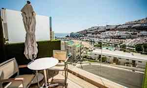 Altan med udsigt over en strand og bjerg dækket med bygninger. (AI-genereret tekst)