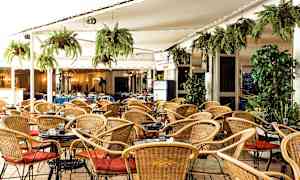 Billede af et udendørsserveringsområde med kurvestole, små borde og grønne hængende planter. (AI-genereret tekst)