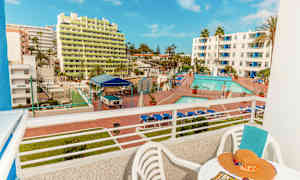 Udsigt fra en balkon, der viser et hotelområde med pool og liggestole samt bygninger i baggrunden. (AI-genereret tekst)