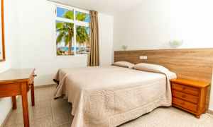 Et hotelværelse med to enkeltsenge beklædt med beige sengetæpper. En væg har et vindue med udsigt til en palme og havet. (AI-genereret tekst)