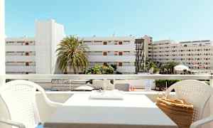 Udsigt fra en altan med siddegruppe, bygninger og palmer synlige i baggrunden under en klar blå himmel. (AI-genereret tekst)