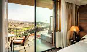 Et hotelværelse med udsigt gennem en glasdør til en altan med stole og et fjernt landskab udenfor. (AI-genereret tekst)