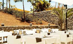 Udendørs restaurant med opstillede borde og stole, omgivet af stenmur og palmer. (AI-genereret tekst)