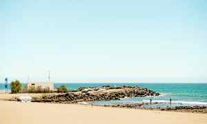 Sandstrand med udsigt over et klippeområde og en lille bådehavn ved havet. (AI-genereret tekst)