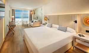 Et hotelværelse med en stor seng, sofa og udsigt over havet gennem vinduer. (AI-genereret tekst)