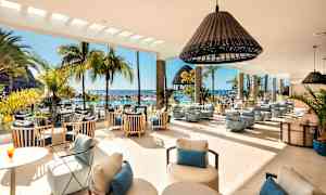 Interiør af en lys dagligstue med moderne møbler, store vinduer og udsigt til et poolområde med palmer og hav. (AI-genereret tekst)