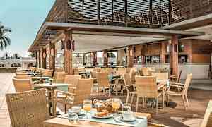 Et udendørs caféområde med træborde, stole og tag, dekoreret med planter og duge, serverer mad og drikke. (AI-genereret tekst)