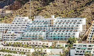 Billede af et hotel med hvid facade og altaner, beliggende ved en klippefyldt skråning. (AI-genereret tekst)