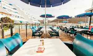 Billede af en udendørs restaurant med borde og blå parasoller. (AI-genereret tekst)