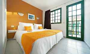 Et soveværelse med en dobbeltseng beklædt med hvide og orange tekstiler, og store vinduer med grønne rammer. (AI-genereret tekst)