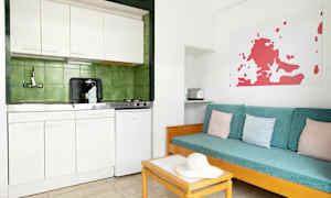 Et enkelt køkken med hvidt interiør og grønne fliser bagved. Ved siden er en sofa med turkise puder og pastelfarvede pudebetræk. (AI-genereret tekst)