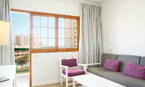 En stue med grå sofa og stole med lilla puder, samt en trødør med glaspartier, der viser en udsigt til bygninger og hav. (AI-genereret tekst)