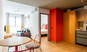 Et moderne indrettet hotelværelse med et bord og stole i forgrunden, en siddegruppe med sofa og et soveværelse synligt gennem en døråbning. Køkkenområdet har skabe og arbejdsområder. (AI-genereret tekst)