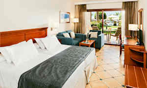 Et hotelværelse med en bred seng, siddeområde og en terrasse med udsigt over en frodig have. (AI-genereret tekst)