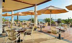 Restaurant med udendørsservering, borde og stole under parasoller med udsigt over havet. (AI-genereret tekst)