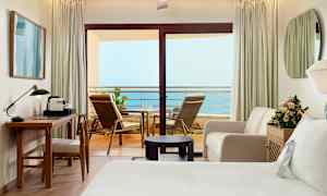 Et hotelværelse med en altan med havudsigt. Værelset er møbleret med skrivebord, stol, sofa, seng og blomster. (AI-genereret tekst)