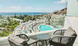 Udsigt fra en altan over et hotel med en stor pool, omgivende palmer og havet i baggrunden. (AI-genereret tekst)