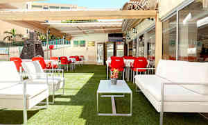 Udendørs servering med hvide sofaer, røde stole og borde, placeret på kunstgræs under en pergola. (AI-genereret tekst)