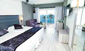 Hotelværelse med dobbeltseng, sofa og havudsigt uden for store vinduer. (AI-genereret tekst)