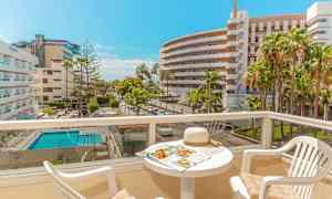 Udsigt over en solrig balkon med bord og stole, hotelkomplekser og pool synlige i baggrunden. (AI-genereret tekst)