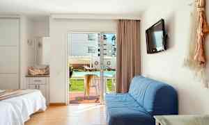 Et hotelværelse med en opredt seng, blå sofa, vægmonteret TV og en glasskydedør, der fører ud til en terrasse med bord og stole. Udsigten viser en udendørspool og liggestole. (AI-genereret tekst)