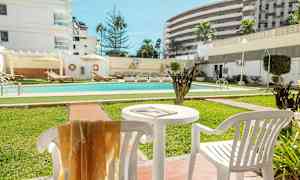 En udendørs pool omgivet af grønne områder og liggestole, med hvide bygninger i baggrunden. (AI-genereret tekst)