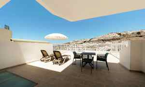 En terrasse med liggestole, parasol og havemøbler med udsigt mod solen og en bjergskråning. (AI-genereret tekst)