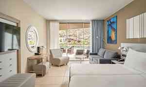 Hotelværelse med to enkeltsenge, sofa og sidde møbler. Glasdøre fører til en terrasse med udsigt. (AI-genereret tekst)