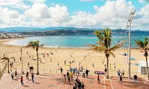 En strand med mennesker, der spiller volleyball og går på strandpromenaden. Havet og bjergene ses i baggrunden. (AI-genereret tekst)