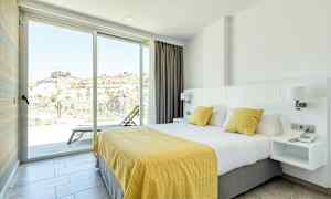 Et hotelværelse med en stor seng dækket med hvidt og gult sengetøj. Værelset har et lyst og rummeligt udseende med glasskydedøre, der leder ud til en balkon med udsigt over et bjergområde. (AI-genereret tekst)