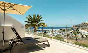 Terrasse med liggestole og parasol, udsigt over lystbådehavn og havet. (AI-genereret tekst)