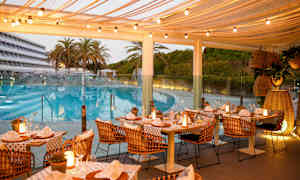 Udendørs restaurant med dækkede borde og lyskæder, beliggende tæt på en pool og omgivet af palmer og grøntområder. (AI-genereret tekst)