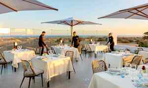 Udendørs servering med dækkede borde og parasoller, med udsigt over sandklitter i baggrunden. (AI-genereret tekst)