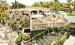 Udendørs restaurant med hvide parasoller og siddepladser, omgivet af grønne områder og palmer. (AI-genereret tekst)