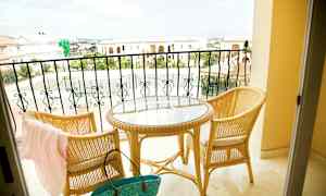 To rottingstole og et rundt bord med glasplade står på en altan med udsigt over stenbygninger og et tennisområde. (AI-genereret tekst)