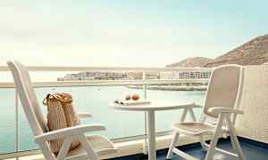 En balkon med udsigt over en havbugt, inklusive to stole og et bord med en bog og briller. (AI-genereret tekst)