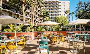Billedet viser en udendørs restaurant med farverige stole, borde og parasoller foran et hotel med altaner. (AI-genereret tekst)