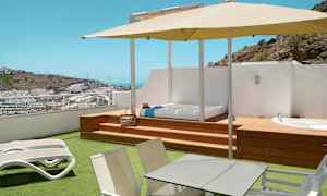 Terrasse med liggestole, bord, parasol og udsigt over et kuperet område med bygninger. (AI-genereret tekst)