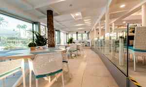 Interiøret i en restaurant med hvide stole og borde, glasvægge og udsigt til udendørs grønne områder. (AI-genereret tekst)