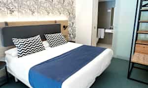 Et moderne hotelværelse med dobbeltseng, blåt sengetæppe og to mønstrede puder. (AI-genereret tekst)